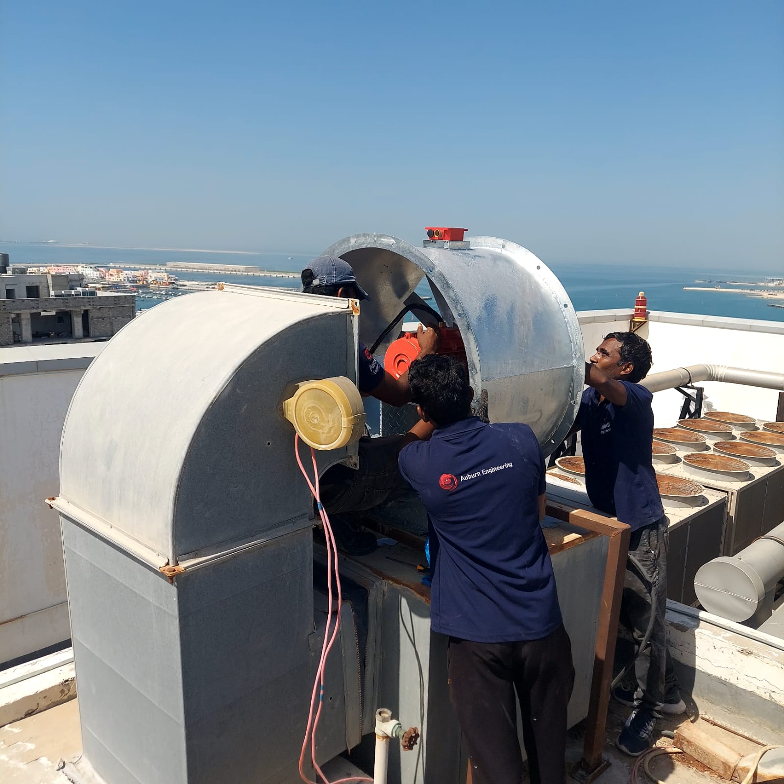 Two technicians install a large rooftop ventilation fan above Doha's coastline.